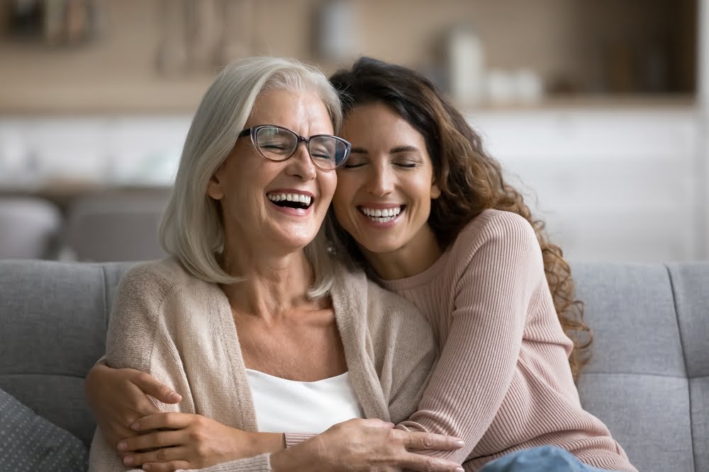 An older woman and a younger woman sit together on a couch, sharing a joyful moment as they laugh and embrace each other - Optometrist Costa Mesa An older woman and a younger woman sit together on a couch, sharing a joyful moment as they laugh and embrace each other - Optometrist Costa Mesa