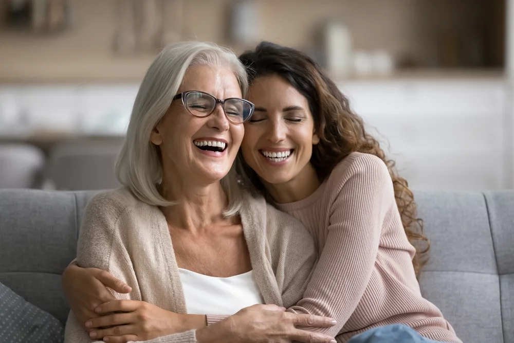 Older woman in glasses laughing with a younger woman on a couch, illustrating the multigenerational family eye care provided at One EyeCare in Costa Mesa 