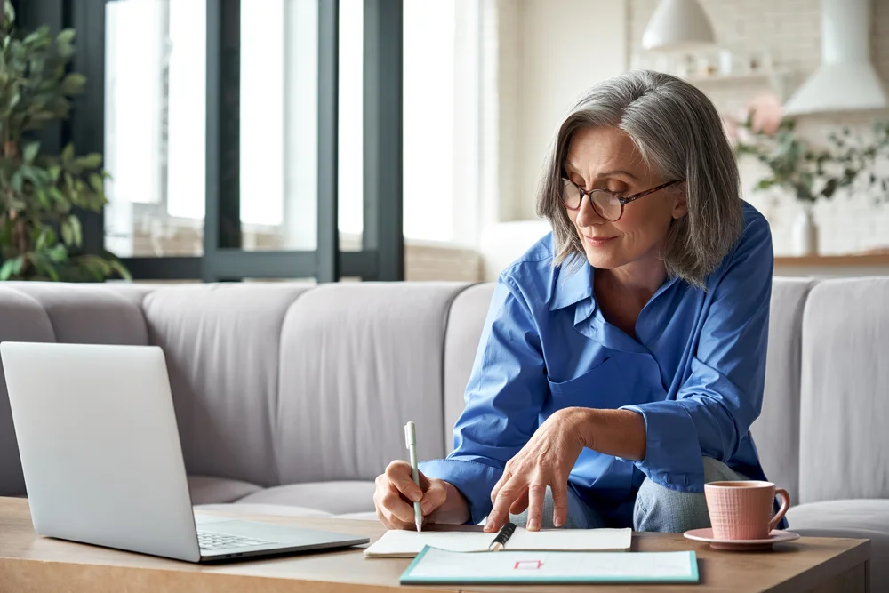 Mature woman wearing glasses and writing notes near a laptop, representing older adult patients who rely on One EyeCare for comprehensive eye care in Costa Mesa 