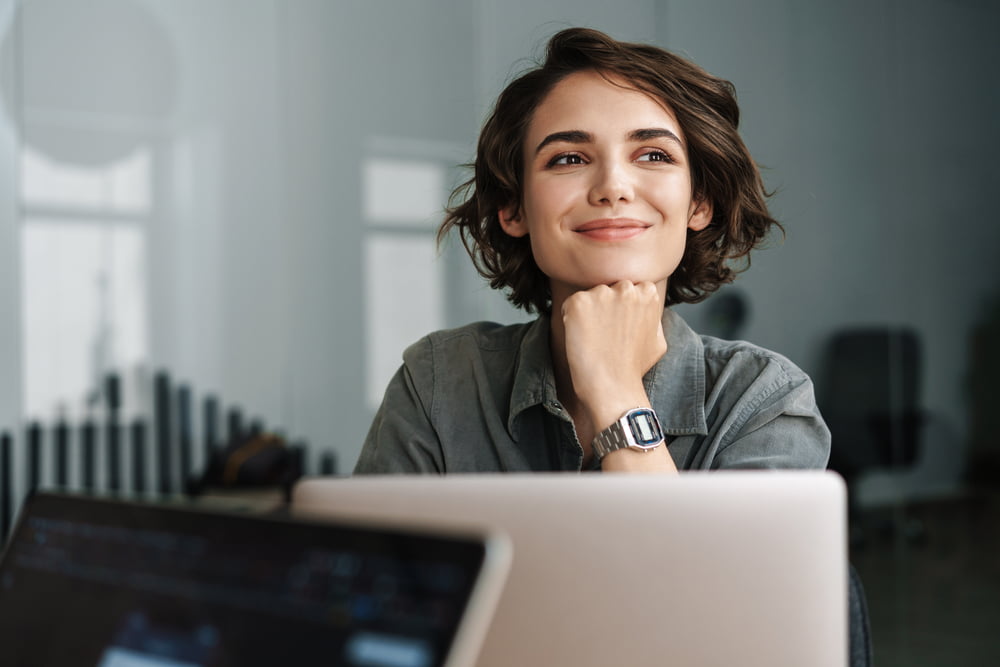 A woman with short hair, wearing a watch, smiles thoughtfully while sitting at a desk in front of a laptop - Optometrist Costa Mesa A woman with short hair, wearing a watch, smiles thoughtfully while sitting at a desk in front of a laptop - Optometrist Costa Mesa