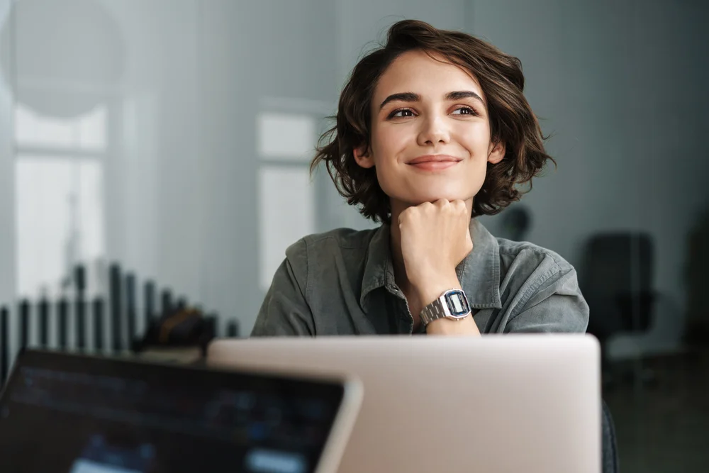 Happy woman sitting at a laptop and looking up with a smile, illustrating the comfort and confidence that comes from personalized eye care at One EyeCare in Costa Mesa