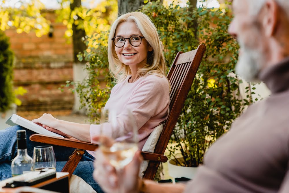 A woman wearing glasses smiles while sitting outdoors with a book, engaging in conversation with another person who is holding a glass of wine - Optometrist Costa Mesa  A woman wearing glasses smiles while sitting outdoors with a book, engaging in conversation with another person who is holding a glass of wine - Optometrist Costa Mesa
