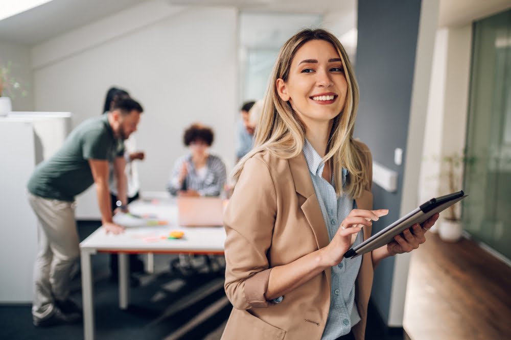 A woman smiles while holding a tablet in an office environment, with colleagues collaborating in the background - Optometrist Costa Mesa A woman smiles while holding a tablet in an office environment, with colleagues collaborating in the background - Optometrist Costa Mesa