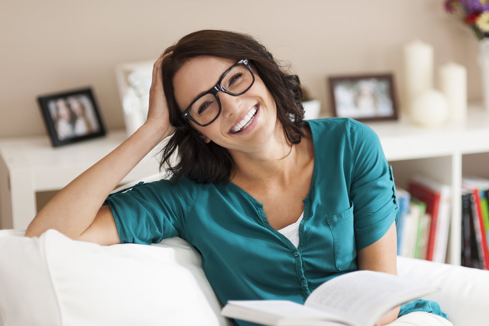 A woman with glasses smiles warmly while sitting on a couch, holding an open book in her hand - Optometrist Costa Mesa A woman with glasses smiles warmly while sitting on a couch, holding an open book in her hand - Optometrist Costa Mesa