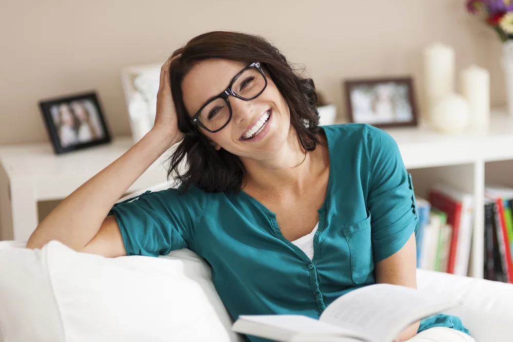 Woman in black-framed glasses smiling while reading at home, showing how well-fitted prescription eyewear supports everyday comfort and clear vision 