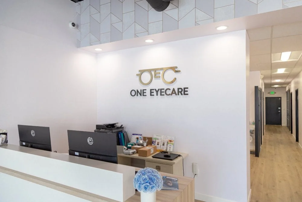 One EyeCare reception desk with the OEC logo on the wall and a clean modern hallway leading to exam rooms, showing the welcoming office environment at the practice on Bristol Street in Costa Mesa 