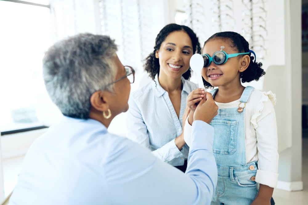 A young girl is trying on optometry testing glasses while a smiling woman, likely her mother, sits beside her, and an optometrist adjusts the frames - Optometrist Costa Mesa A young girl is trying on optometry testing glasses while a smiling woman, likely her mother, sits beside her, and an optometrist adjusts the frames - Optometrist Costa Mesa