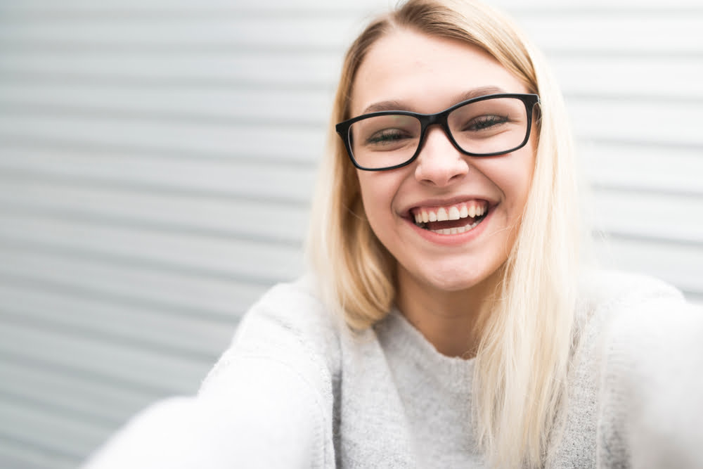 A smiling woman with blonde hair and black glasses, wearing a light sweater, takes a selfie in front of a light-colored background - Optometrist Costa Mesa A smiling woman with blonde hair and black glasses, wearing a light sweater, takes a selfie in front of a light-colored background - Optometrist Costa Mesa