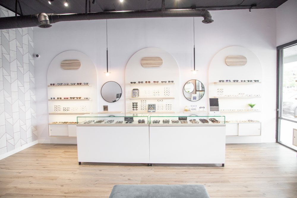 A bright, modern optometry store interior with white walls, wooden floors, and neatly displayed eyewear on shelves and in glass cases, with two round mirrors on the back wall - Optometrist Costa Mesa A bright, modern optometry store interior with white walls, wooden floors, and neatly displayed eyewear on shelves and in glass cases, with two round mirrors on the back wall - Optometrist Costa Mesa