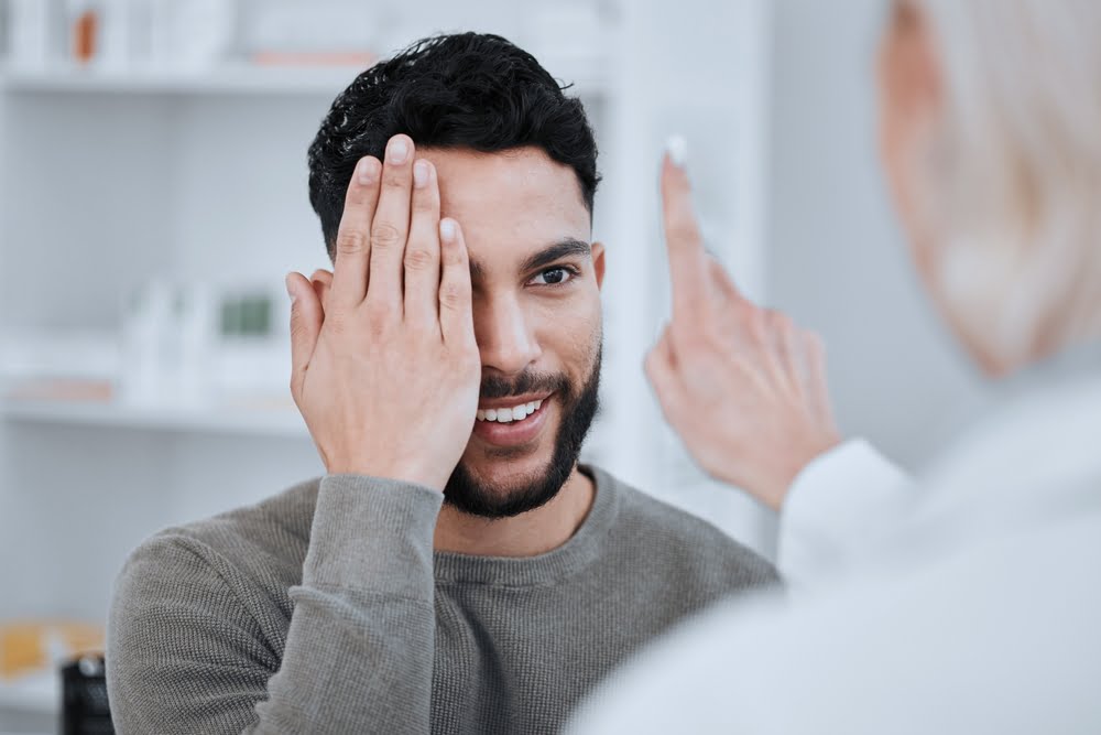 A man covers one eye with his hand while undergoing an eye examination with an optometrist, who is pointing in front of him - Optometrist Costa Mesa A man covers one eye with his hand while undergoing an eye examination with an optometrist, who is pointing in front of him - Optometrist Costa Mesa