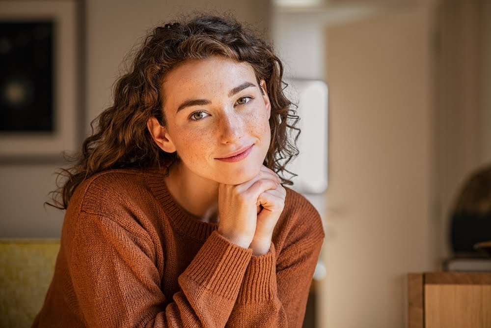 A woman with curly hair and freckles rests her chin on her hands, smiling softly while wearing a cozy brown sweater - Optometrist Costa Mesa A woman with curly hair and freckles rests her chin on her hands, smiling softly while wearing a cozy brown sweater - Optometrist Costa Mesa