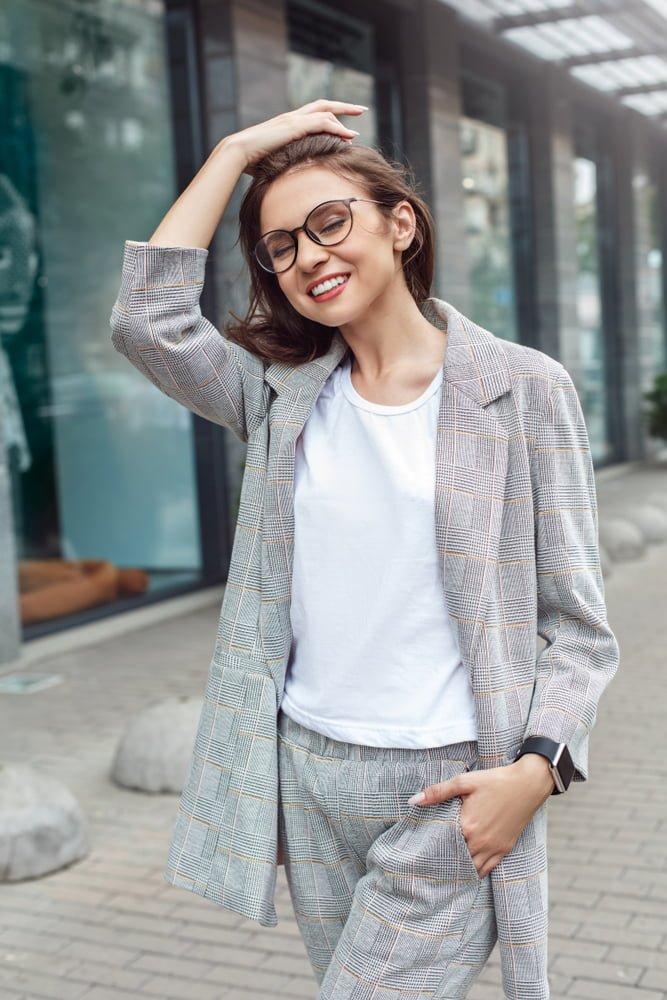A woman smiles confidently outdoors while adjusting her glasses, wearing a plaid suit and white shirt - Optometrist Costa Mesa A woman smiles confidently outdoors while adjusting her glasses, wearing a plaid suit and white shirt - Optometrist Costa Mesa